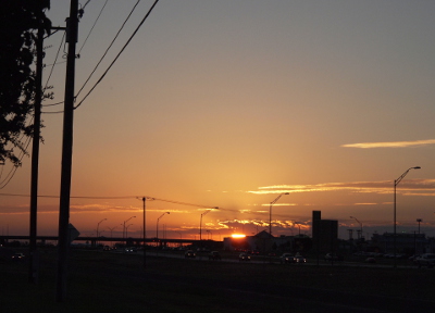 [The bright yellow-orange orb of the sun is just above the horizon, but its light is partially blocked by the buildings in the distance and the cloud bank above the horizon. The upper part of the sky is cloud-free and thus rays eminating from the sun can be seen.]
